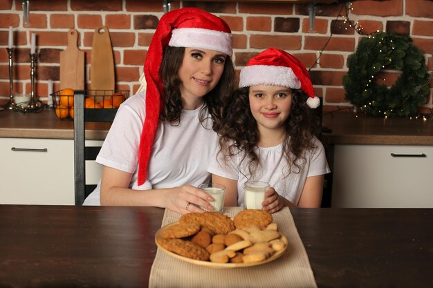 mamma e figlia con i cappelli di babbo natale in cucina che tengono il latte sul tavolo biscotti di farina d'avena