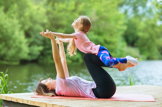 Mamma e figlia eseguono un esercizio di yoga sulla riva del fiume in una calda giornata di sole