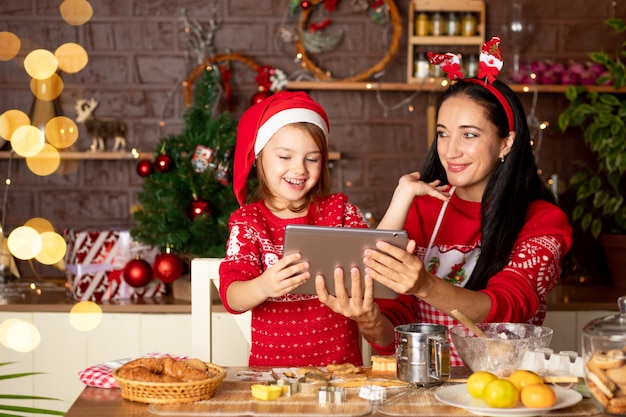 Mamma e figlia stanno parlando tramite collegamento video, passando gli auguri su un tablet in una cucina buia con un albero di Natale per Capodanno o Natale, sorridendo e divertendosi insieme in un cappello di Babbo Natale