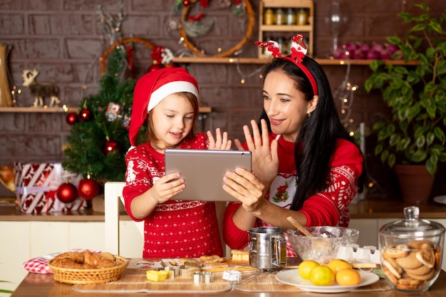 Mamma e figlia stanno parlando tramite collegamento video, passando gli auguri su un tablet in una cucina buia con un albero di Natale per Capodanno o Natale, sorridendo e divertendosi insieme in un cappello di Babbo Natale