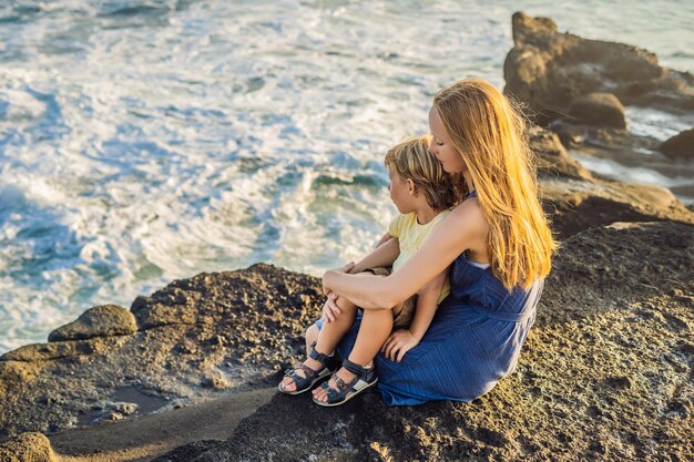 Mamma e figlio sono seduti su una roccia e guardano il mare. Turisti di viaggio del ritratto - mamma con bambini. Emozioni umane positive, stili di vita attivi. Felice giovane famiglia sulla spiaggia del mare