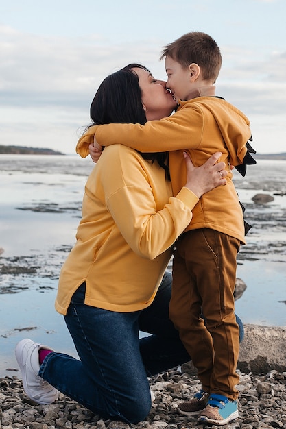 Mamma e figlio sulla riva del fiume. Una bella ragazza con suo figlio in felpa gialla sorride e bacia sinceramente. Ricreazione all'aperto.