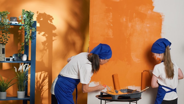 Mamma e ragazza usano la vernice arancione per colorare le pareti interne di casa, divertendosi con il miglioramento della ristrutturazione della casa. Dipingere lo spazio dell'appartamento insieme a pennello e strumenti a rullo. Scatto da treppiede.
