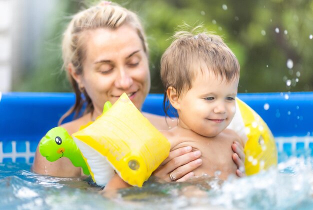 Mamma felice che gioca con il piccolo bambino nudo che ride con bracciali gonfiabili rossi colorati, in piscina con acqua fresca e pulita sullo sfondo del tramonto soleggiato estivo dorato brillante