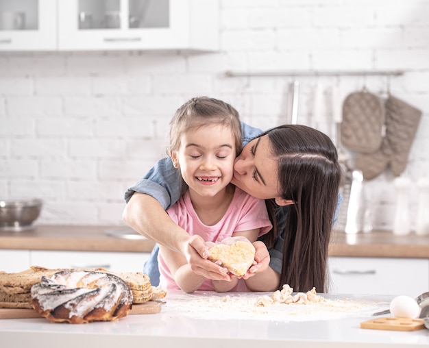 Mamma felice con la figlia che prepara torte fatte in casa sullo sfondo di una cucina leggera.