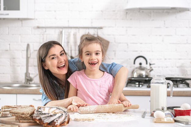 Mamma felice con la figlia che prepara torte fatte in casa sullo sfondo di una cucina leggera.