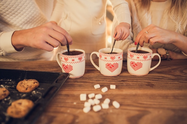 Mamma, papà e figlia di famiglia in maglioni bianchi cucinano e bevono cacao con marshmallow