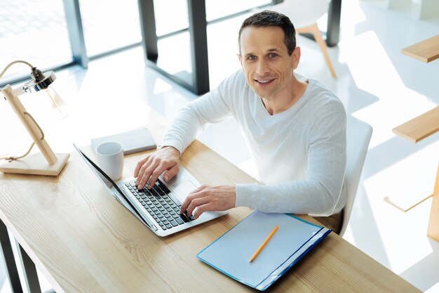 Manager di successo. Uomo intelligente felice attraente che ti guarda ed è di buon umore mentre si lavora al laptop