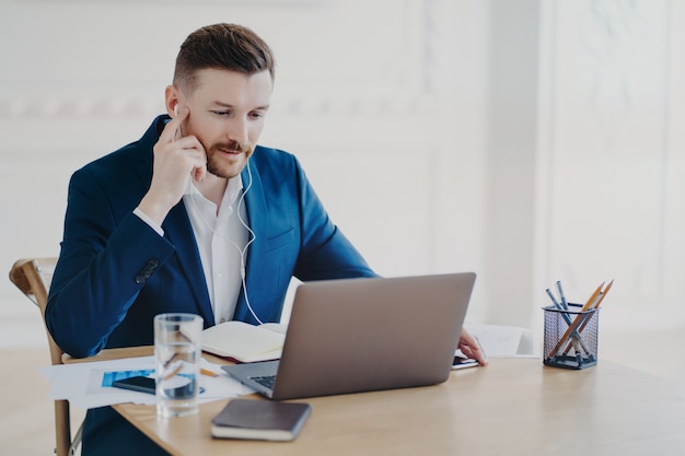 Manager esecutivo di successo che indossa un abito blu formale utilizzando gli auricolari che hanno riunioni online con i dipendenti, l'uomo d'affari è serio mentre è seduto davanti al laptop dietro la scrivania dell'ufficio e parla