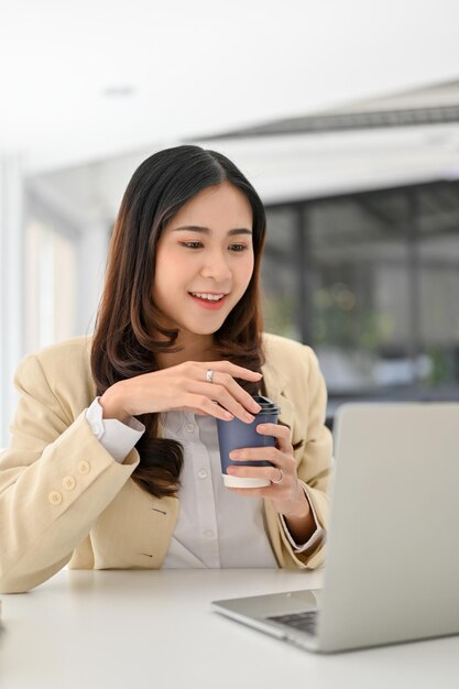 Manager femminile asiatico sorseggiando caffè mentre lavora alle sue attività aziendali sul laptop in ufficio