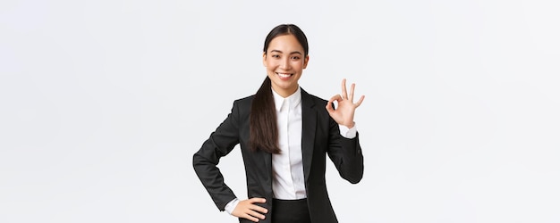 Manager femminile professionale in abito nero che sembra assertivo incoraggiare tutto bene assicurare il lavoro svolto mostrando un gesto corretto e sorridente in approvazione in piedi soddisfatto su sfondo bianco
