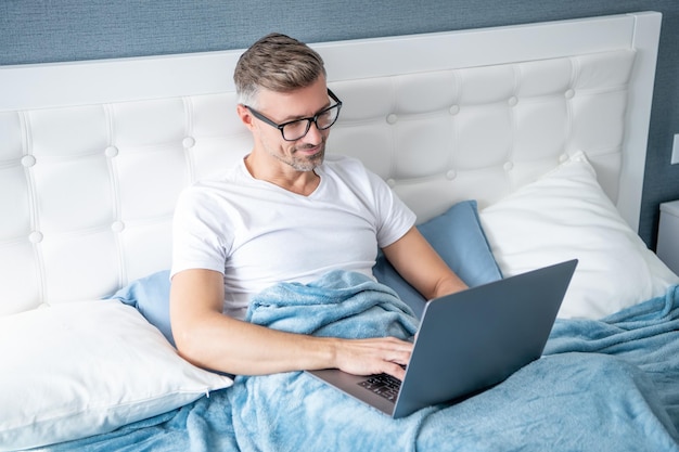 Manager online maturo con gli occhiali che lavora al computer portatile a letto