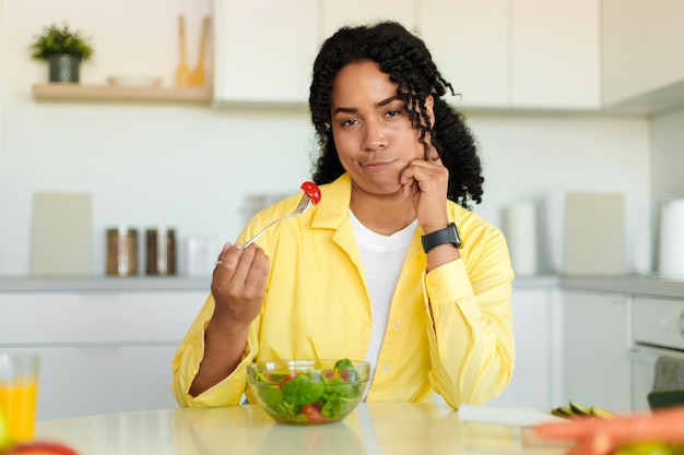 Mancanza di appetito Donna nera infelice che tiene forchetta con pomodoro mentre mangia insalata di verdure seduta in cucina