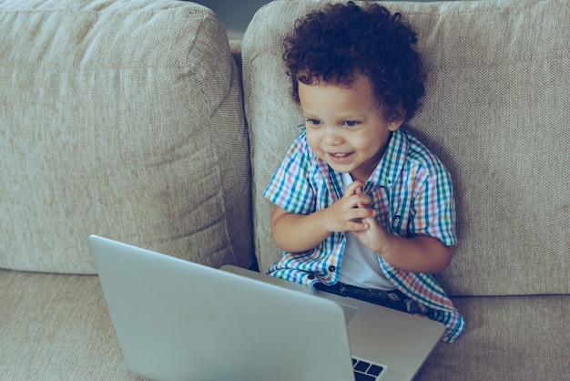 Mandiamo e-mail alla nonna! Veduta dall'alto del piccolo bambino africano che sorride mentre è seduto sul divano di casa con il computer portatile sulle ginocchia