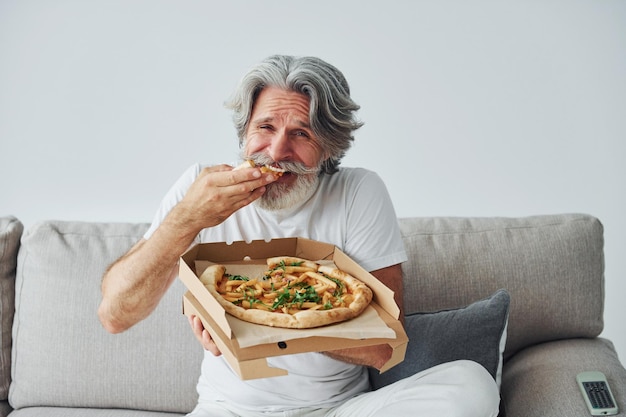 Mangia una deliziosa pizza mentre guardi il programma TV Uomo moderno ed elegante con capelli grigi e barba all'interno