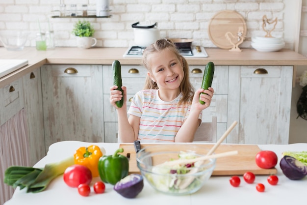 Mangiare sano è divertente. Ragazza carina che cucina insalata di verdure e tiene i cetrioli in cucina, copia spazio