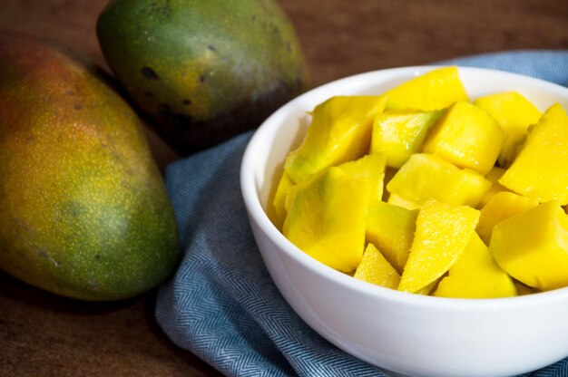 Mango tritato in una ciotola bianca su un panno blu con due frutti interi con la buccia sul lato