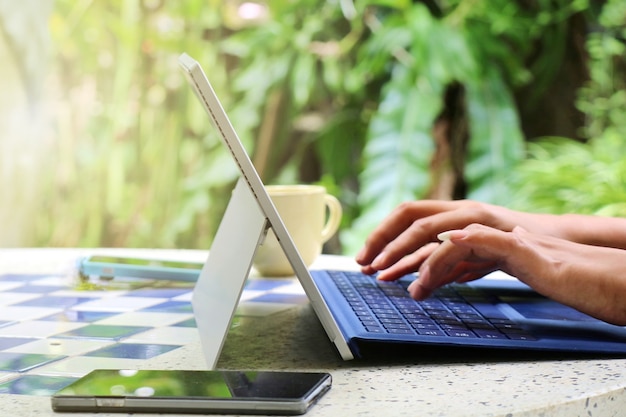 mani che digitano sulla tastiera del laptop con una tazza di caffè e uno smartphone su un tavolo in giardino