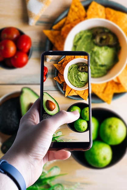 Mani che prendono uno smartphone che prende un'immagine di una ciotola con guacamole. Spazio per il testo. Messa a fuoco selettiva