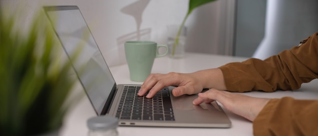 Mani che scrivono sul computer portatile dello schermo in bianco sul tavolo da lavoro semplice bianco con la tazza e le decorazioni