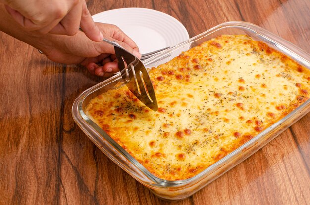 Mani che tagliano le lasagne sul piatto di vetro Composizione del concetto di cottura per cucinare le lasagne su legno.