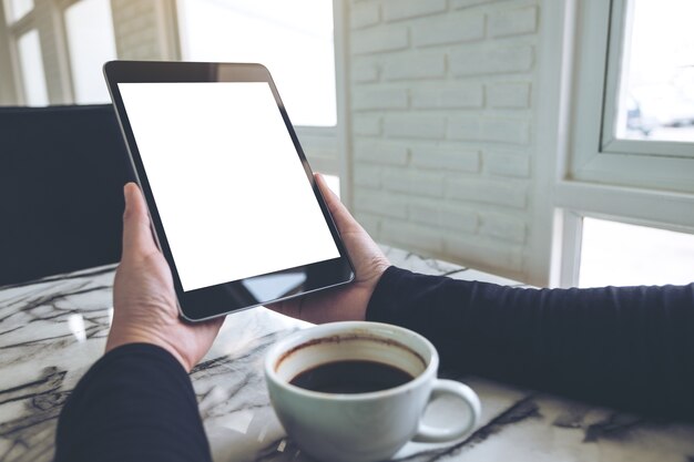 mani che tengono tablet pc nero con schermo bianco vuoto e tazza di caffè sul tavolo nella caffetteria