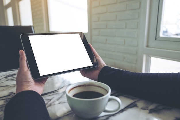 mani che tengono tablet pc nero con schermo bianco vuoto e tazza di caffè sul tavolo nella caffetteria