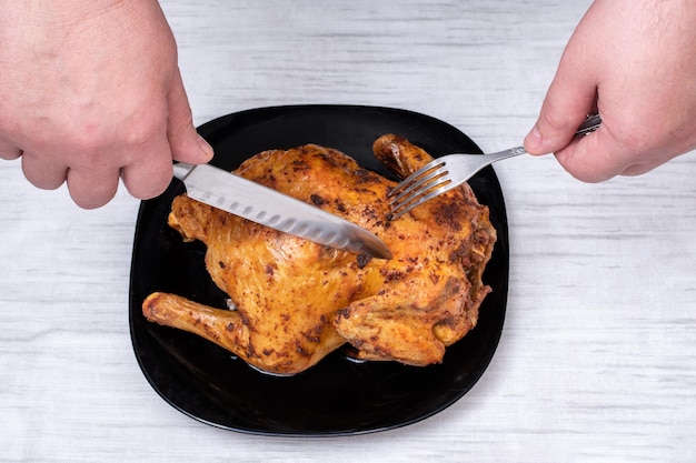 Mani che tengono un coltello e una forchetta per tagliare il pollo fritto su un piatto nero sul tavolo, vista dall'alto. Pollo con una crosta croccante marrone. Pollo fatto in casa. Cibo tradizionale di Natale.