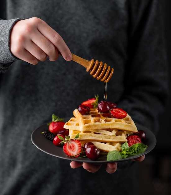 Mani che tengono un piatto di waffle belga con fragole fresche bacche foglie di menta e bagnato di miele su uno sfondo scuro
