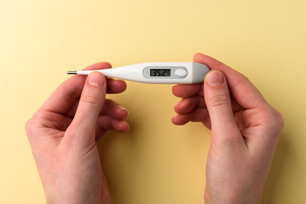 Mani che tengono un termometro elettronico con temperatura normale sul display su sfondo giallo.