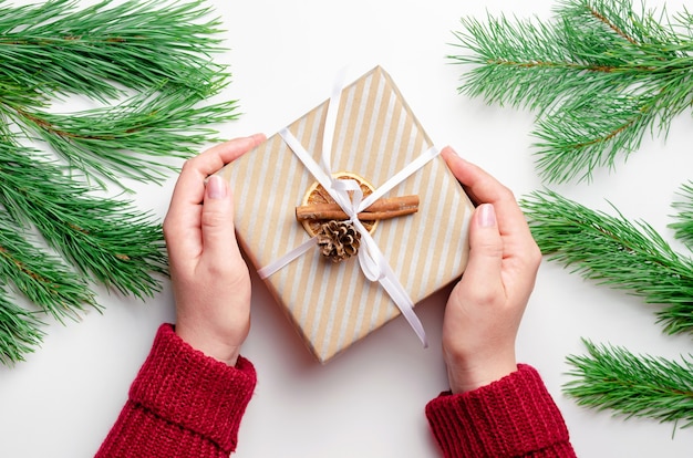 Mani che tengono una scatola regalo di natale decorata su sfondo bianco con rami di pino