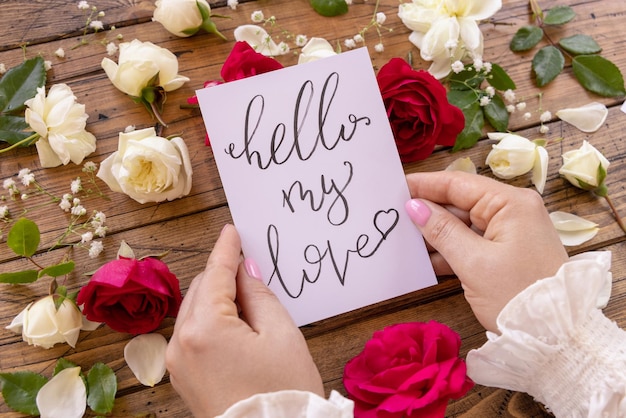 Mani con carta scritta a mano HELLO MY LOVE circondate da rose rosse e crema si chiudono su un tavolo di legno. Dichiarazione romantica femminile d'amore vicino ai fiori, concetto di San Valentino