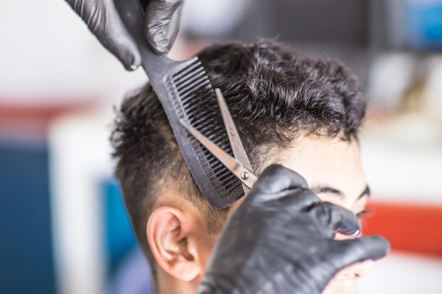 Mani con guanti di un barbiere che tagliano i capelli di un cliente latino usando un pettine e forbici