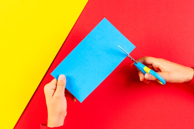 Mani del bambino che tagliano carta di colore blu con le forbici su sfondo di carta rossa e gialla