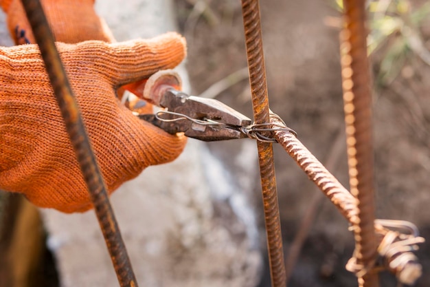 Mani del costruttore in guanti arancioni, torcendo l'armatura con filo usando una pinza