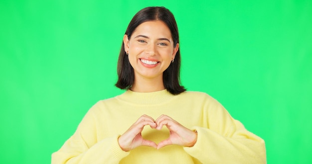 Mani del cuore e volto di donna felice sullo studio con schermo verde o sorriso su sfondo a colori Ritratto del modello femminile e forma del dito dell'amore grazie o supporto del segno emoji della pace o cura della gentilezza