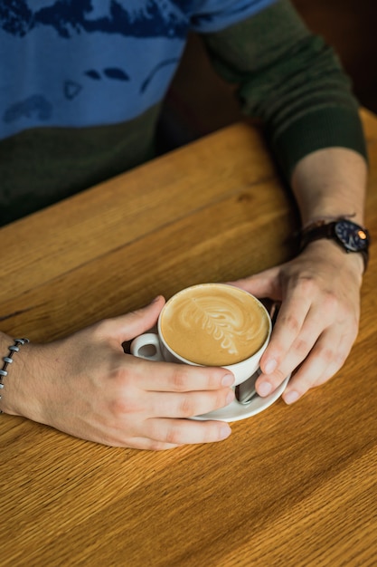 Mani del maschio che tengono una tazza di caffè cappuccino, tavolo in legno