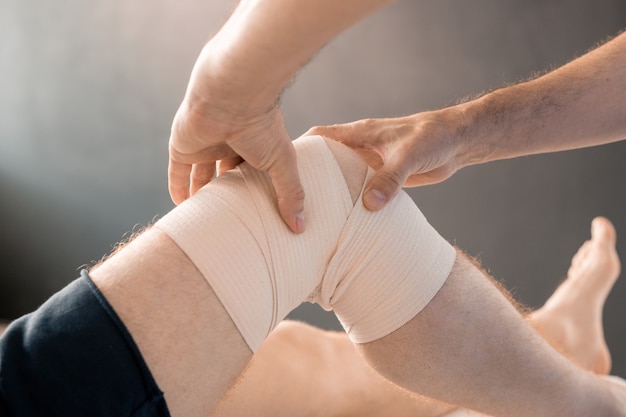 Mani del medico contemporaneo che massaggiano il ginocchio di un giovane paziente di sesso maschile avvolto con una fasciatura flessibile durante il corso di riabilitazione