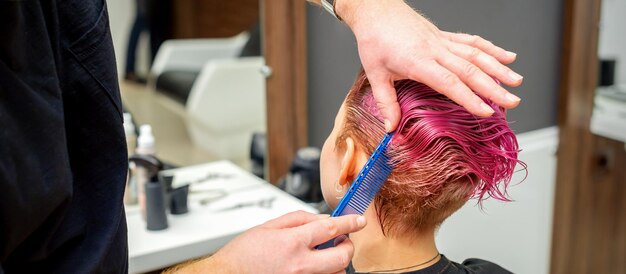 Mani del parrucchiere che pettinano i capelli rendendo l'acconciatura rosa corta per una giovane donna caucasica in un salone di bellezza.