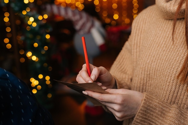 Mani del primo piano della giovane donna che scrive la lettera di Natale a Babbo Natale sull'albero di natale del fondo