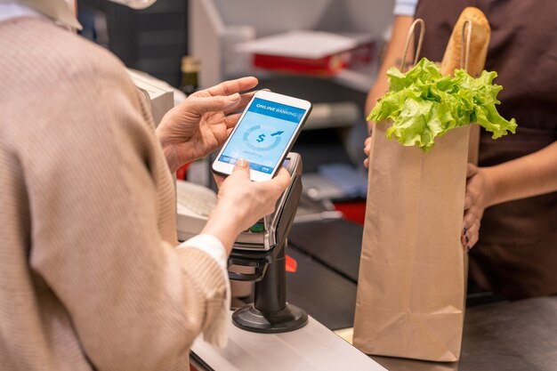 Mani dell'acquirente femminile maturo con lo smartphone sulla macchina di pagamento andando a pagare per i prodotti alimentari nel supermercato tramite registratore di cassa