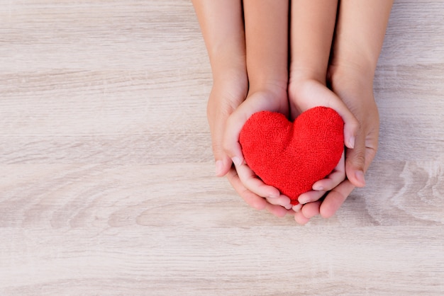 Mani dell&#39;adulto e del bambino che tengono cuore rosso fatto a mano su fondo di legno.