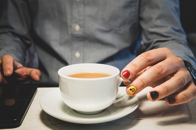 Mani dell'uomo che tengono una tazza di tè e usa uno smartphone sul tavolo di legno. Un uomo con le unghie dipinte. Progettazione di unghie maschili. manicure da uomo.