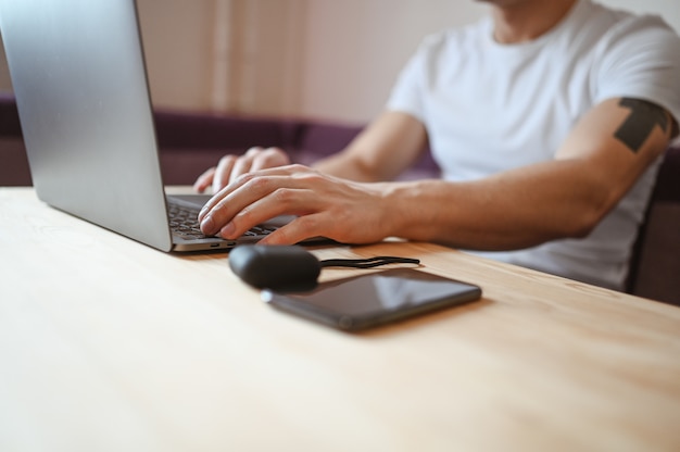 Mani dell'uomo irriconoscibile con lo smartphone e le cuffie che lavorano ad un computer portatile durante l'isolamento del coronavirus