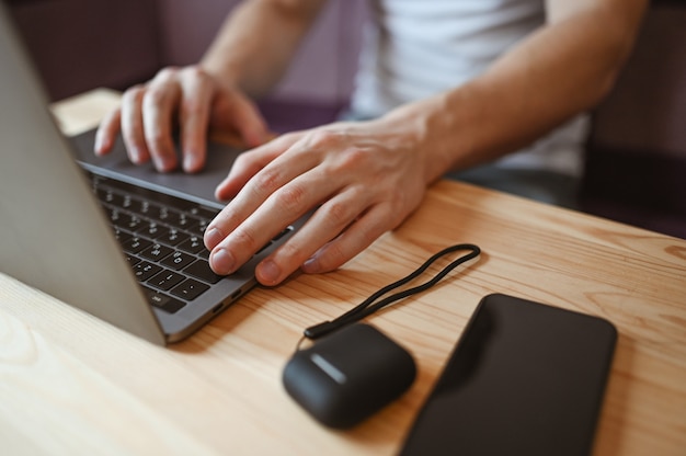 Mani dell'uomo irriconoscibile con lo smartphone e le cuffie che lavorano ad un computer portatile durante l'isolamento del coronavirus