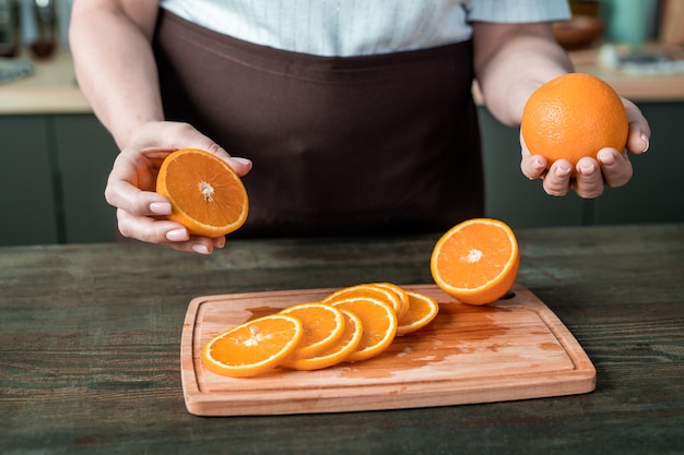 Mani della casalinga contemporanea in grembiule in piedi dal tavolo della cucina e che tiene le arance fresche sul bordo di legno con fette succose