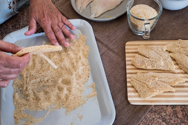 Mani della donna anziana che preparano i filetti di pesce impanati con le uova Freschezza in tavola con tagliere in legno