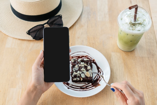 Mani della donna asiatica utilizza lo smartphone mentre si mangia la torta brownie con latte Matcha ghiacciato