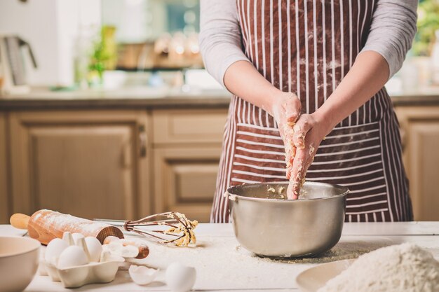 Mani della donna che impastano la pasta in una ciotola di ferro. Una donna con un grembiule a righe sta cucinando in cucina