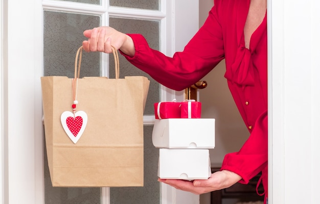 Mani della donna che tengono il sacchetto della spesa con cuori rossi di San Valentino e contenitori di regali vicino alla porta bianca.